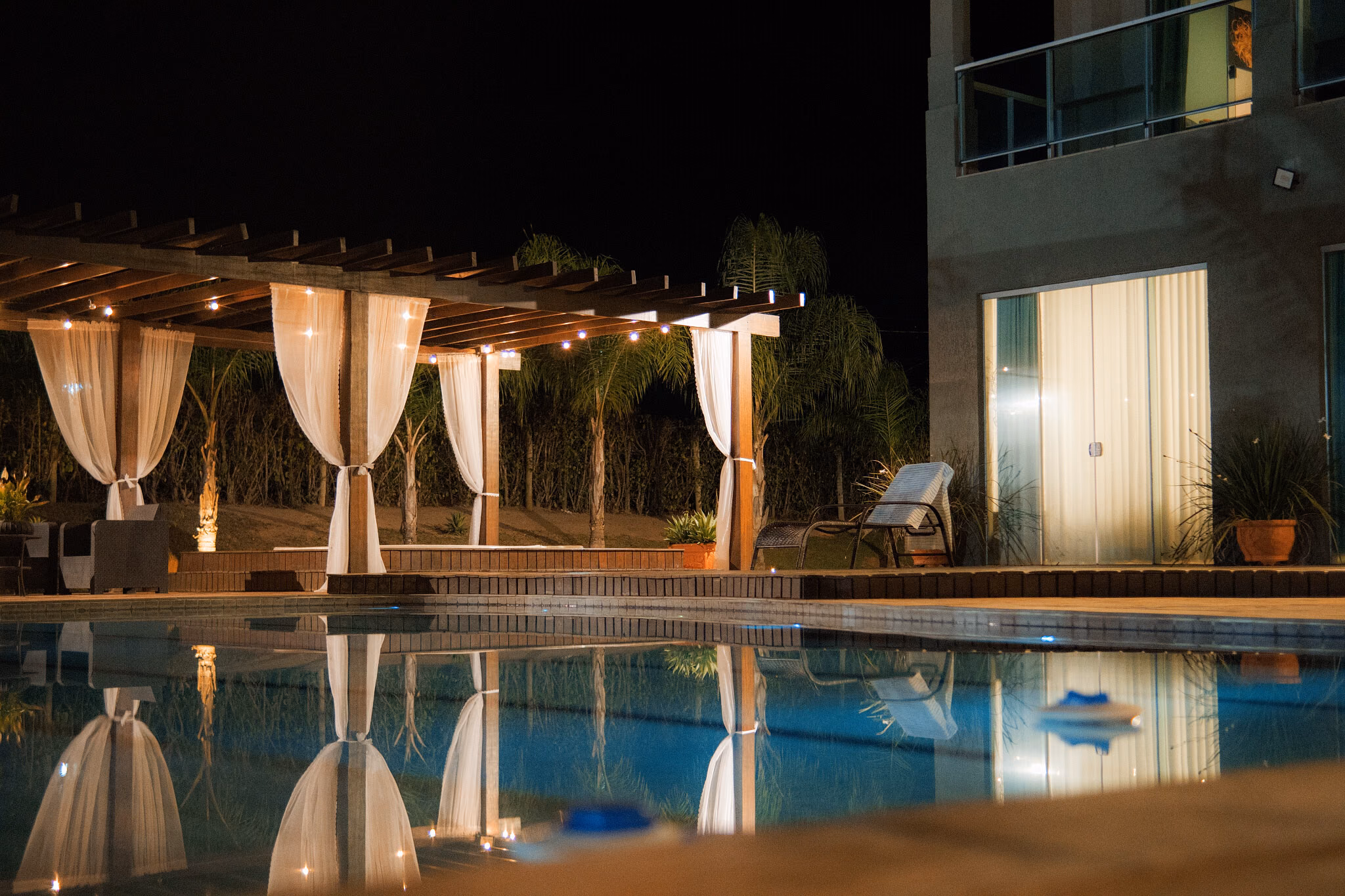 Pergolado à beira da piscina à noite, com voile branco iluminado e reflexo na água